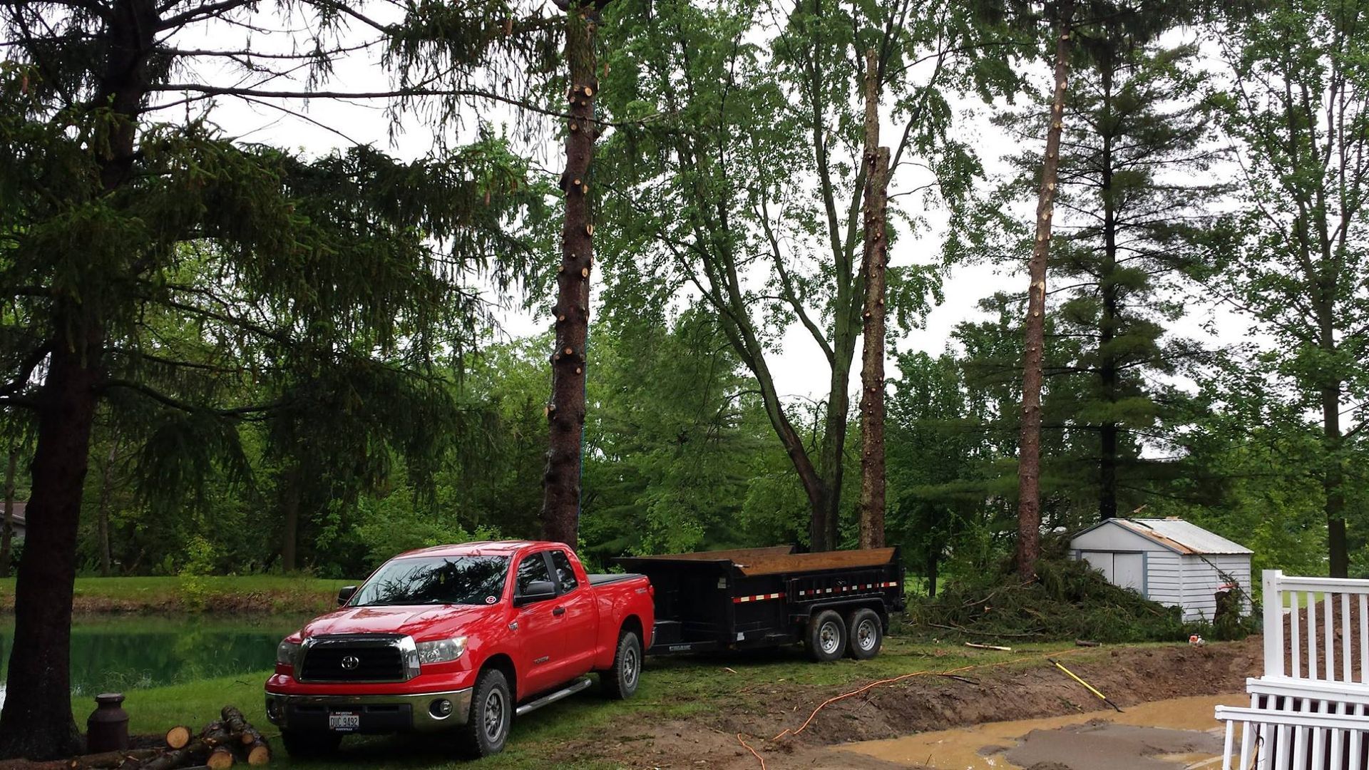 A red pickup truck pulling a dump trailer is parked on a grassy lawn next to tall trees and a small white shed.