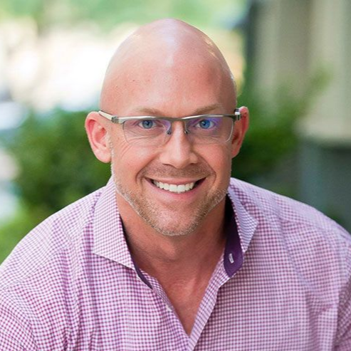Smiling bald man wearing glasses and a pink checkered shirt, posed outdoors against a blurred green background
