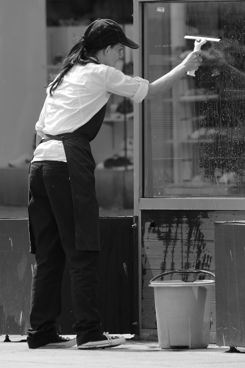 Person cleaning a window with a squeegee, wearing an apron and cap. A bucket sits nearby.