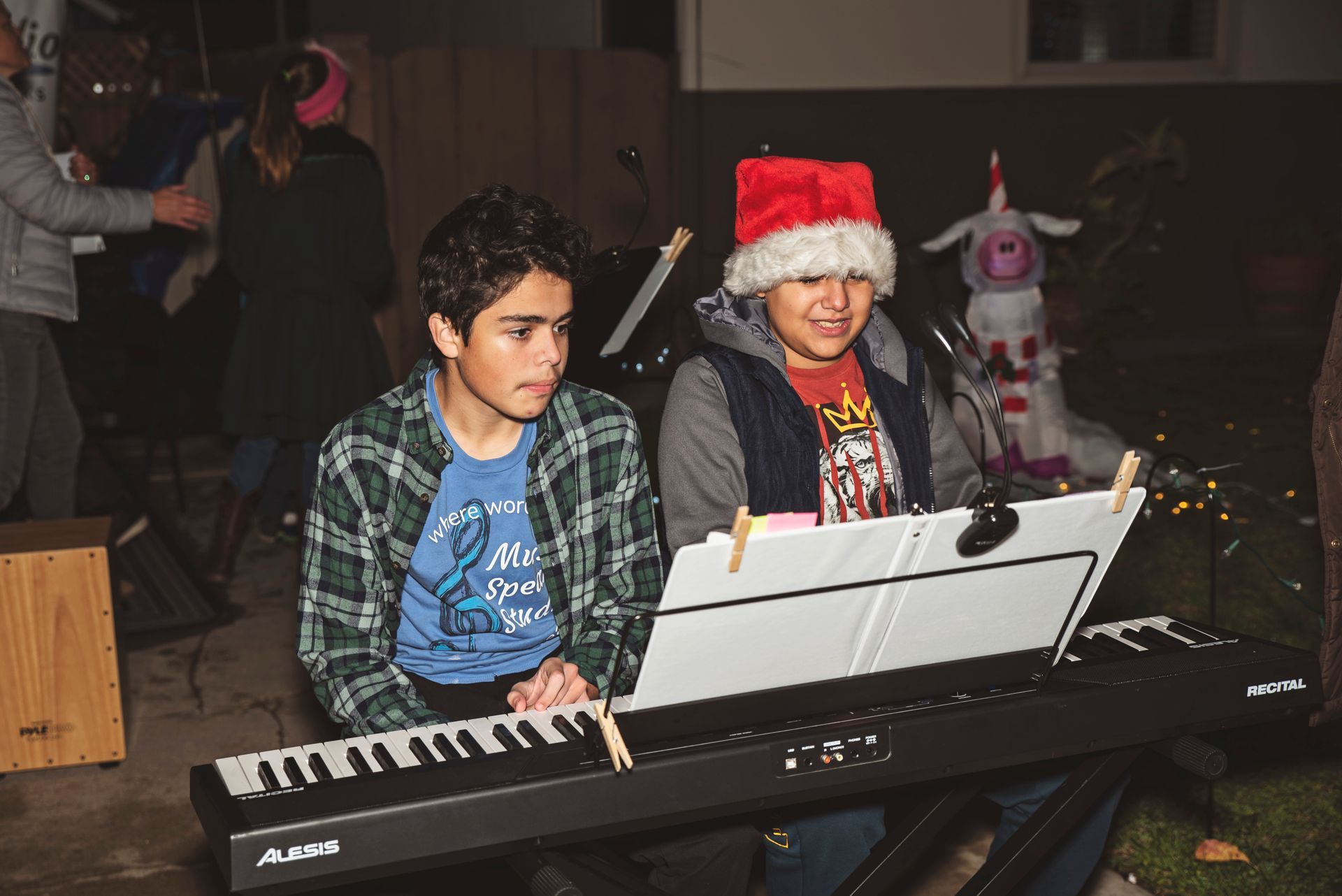 Two boys are sitting at a piano and one of them is wearing a santa hat.