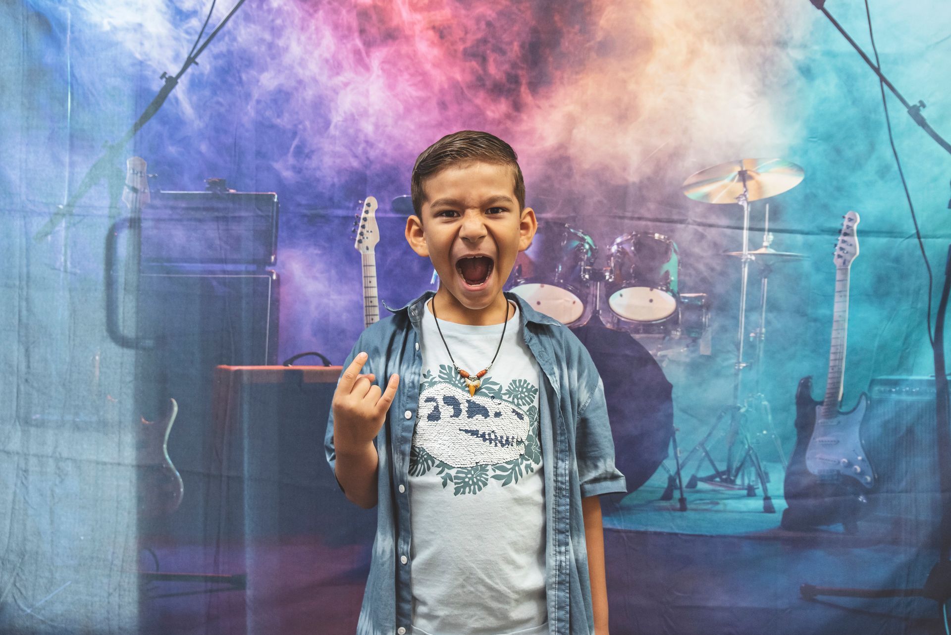 A young boy is making a rock and roll sign in front of a drum set.