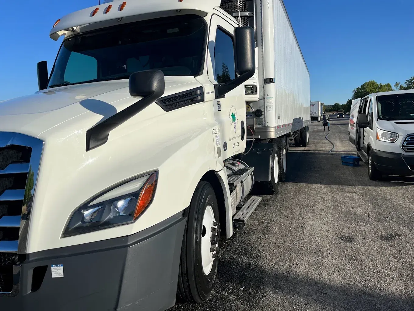White semi-truck parked on asphalt, side view. Another white van is nearby. Blue sky.