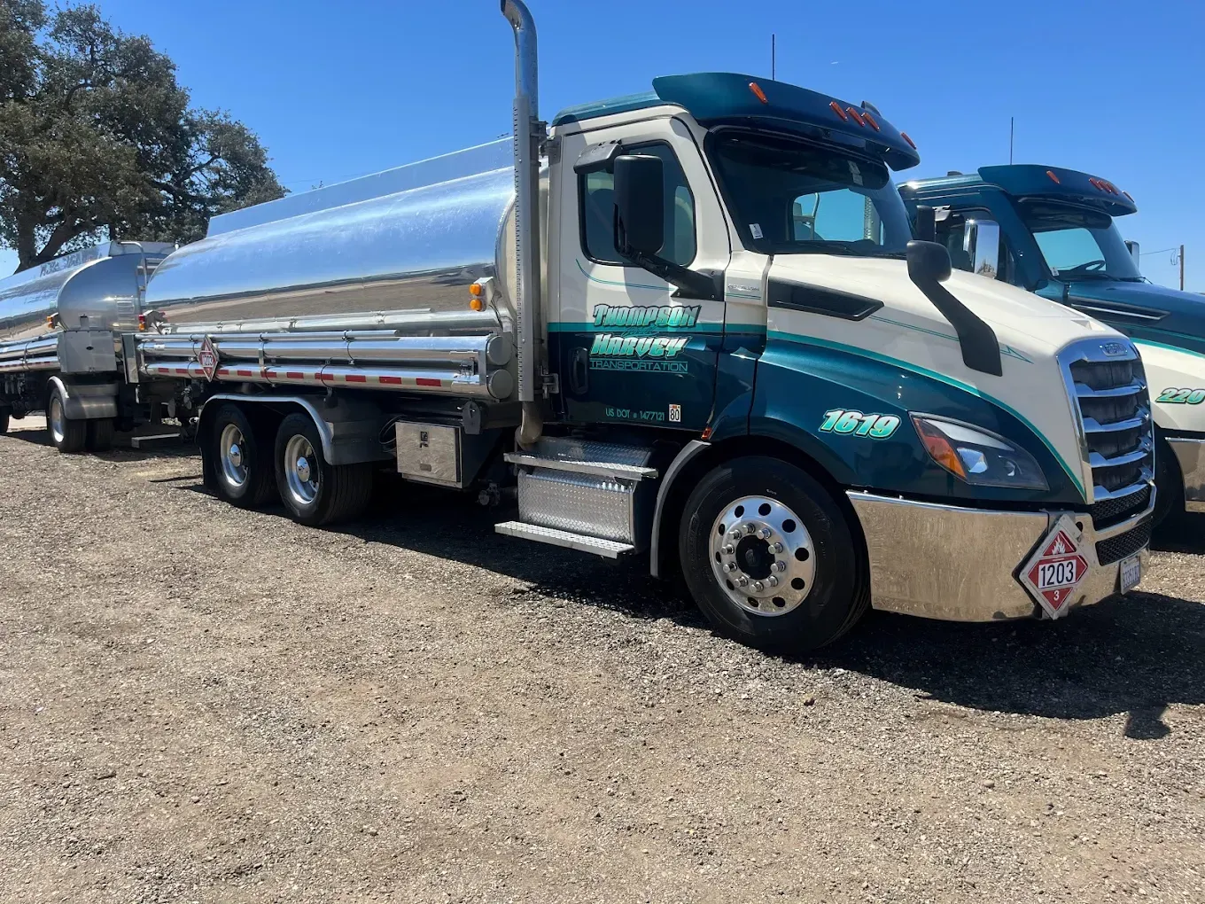 Fuel tanker truck, cream and teal, parked on gravel in a sunny lot.