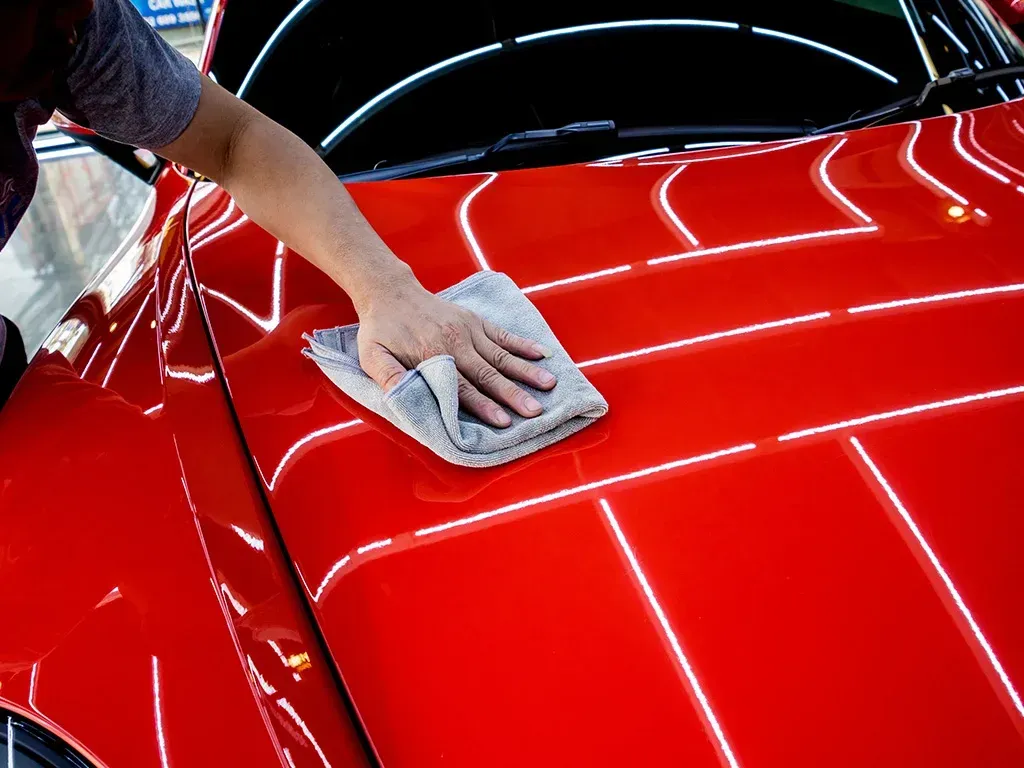 Person polishing a shiny red car hood with a gray microfiber cloth.
