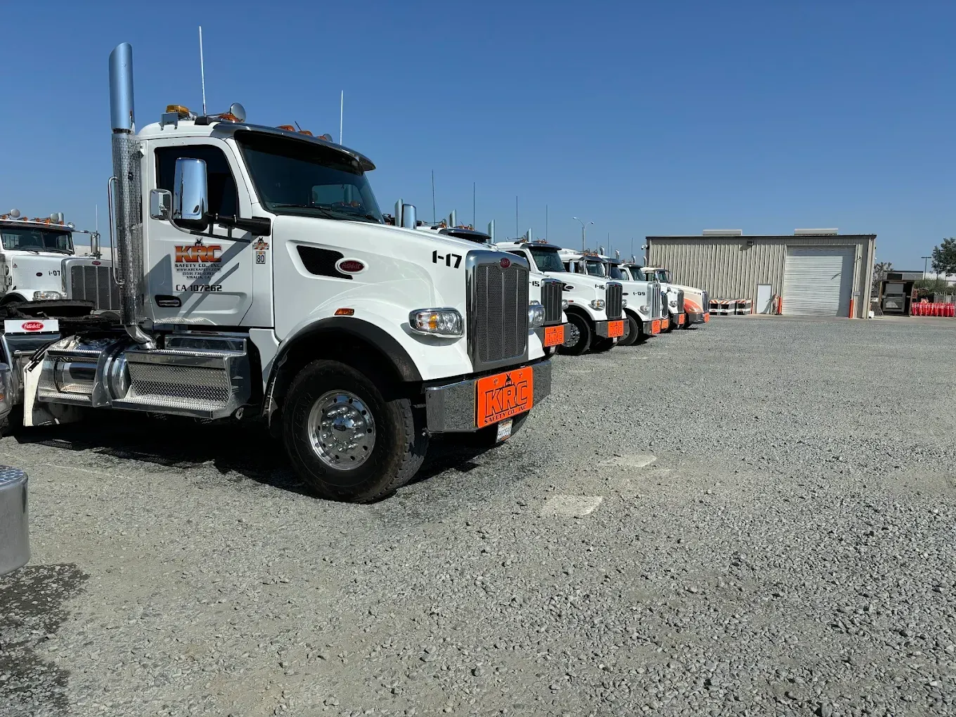 White semi-trucks parked in a gravel lot on a sunny day. A small building is in the background.