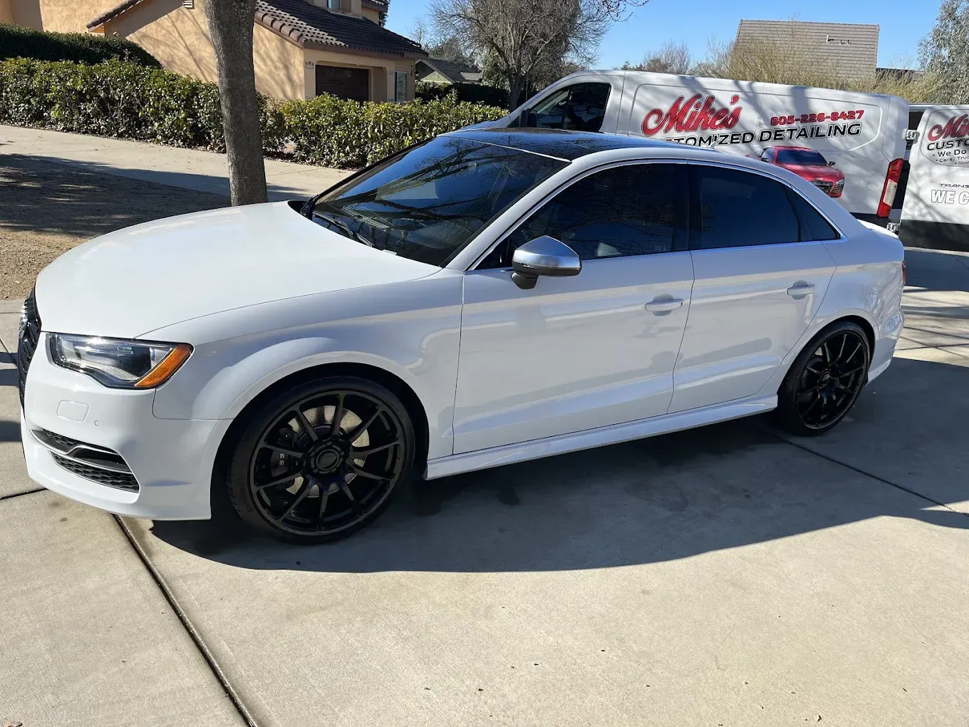 White Audi sedan with tinted windows, parked on a driveway, with a black rimmed wheels, and a commercial van in the background.