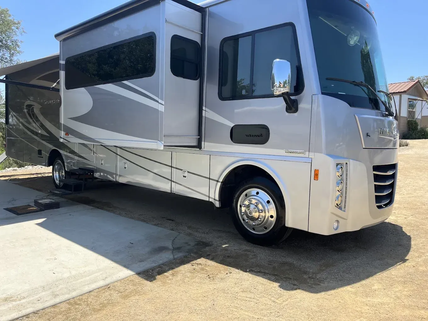 Silver RV parked on a gravel driveway with awning extended on a sunny day.