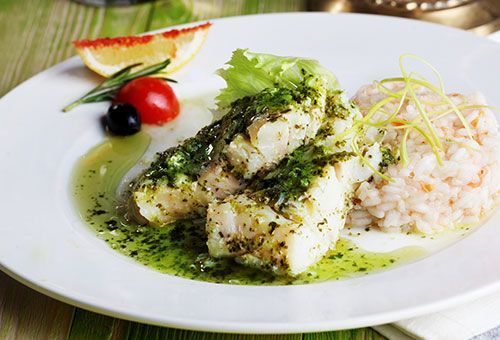 Plate of baked fish with green sauce, rice, lemon, tomato, and olive.