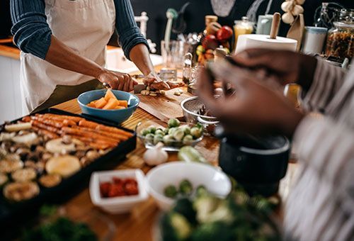 People preparing a meal in a kitchen, chopping vegetables, with roasted vegetables on a tray and other ingredients.