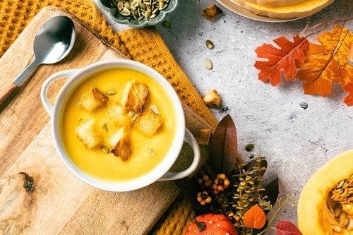 A bowl of creamy pumpkin soup with croutons, autumn leaves, and pumpkin seeds on a wooden board.