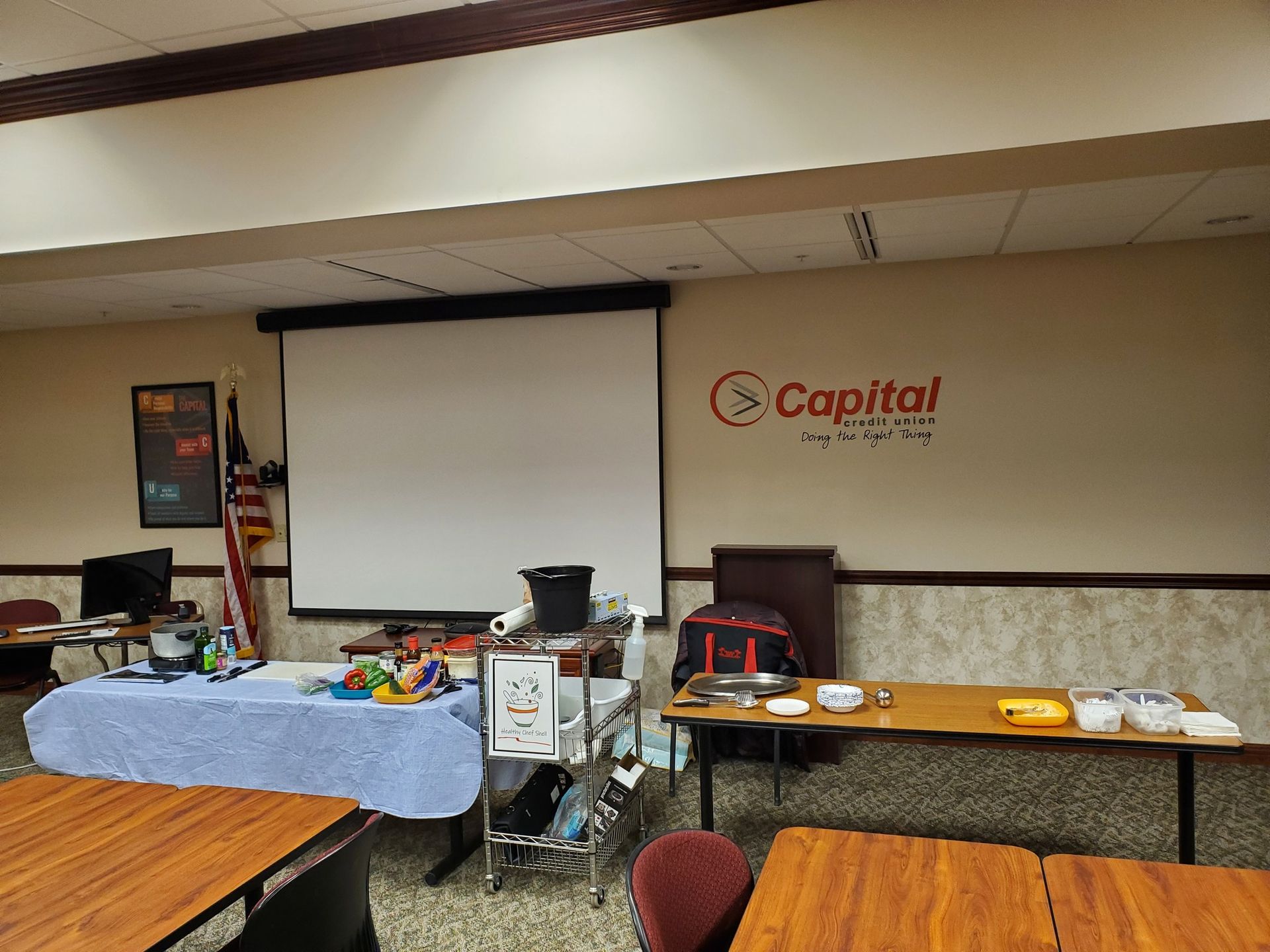 A room with tables , chairs , a projector screen and a capital credit union sign on the wall.