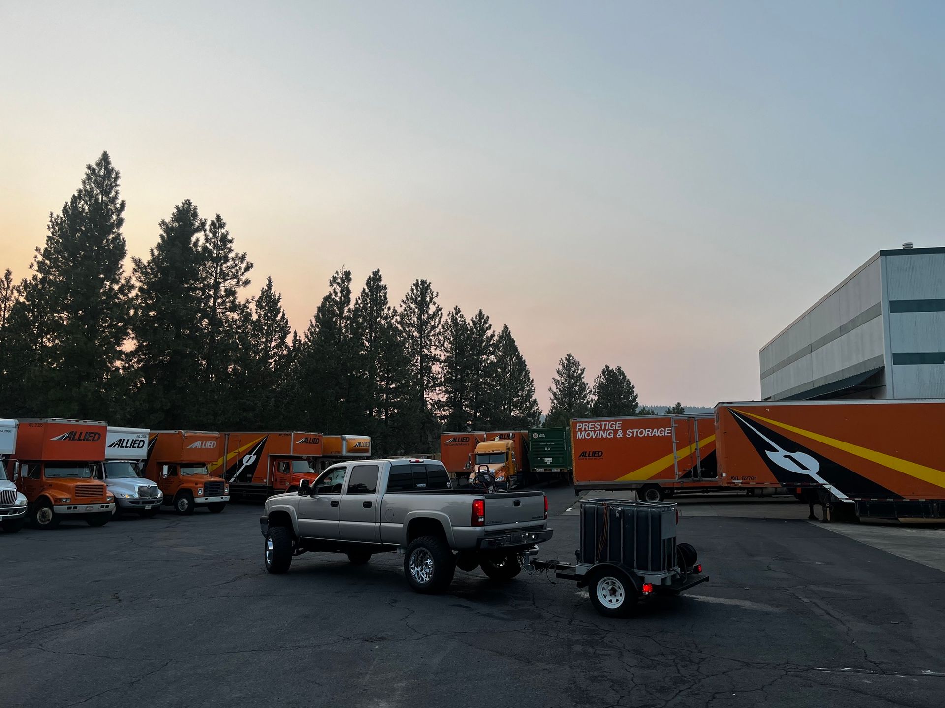 A silver pickup truck towing a small trailer in a parking lot filled with orange moving trucks.