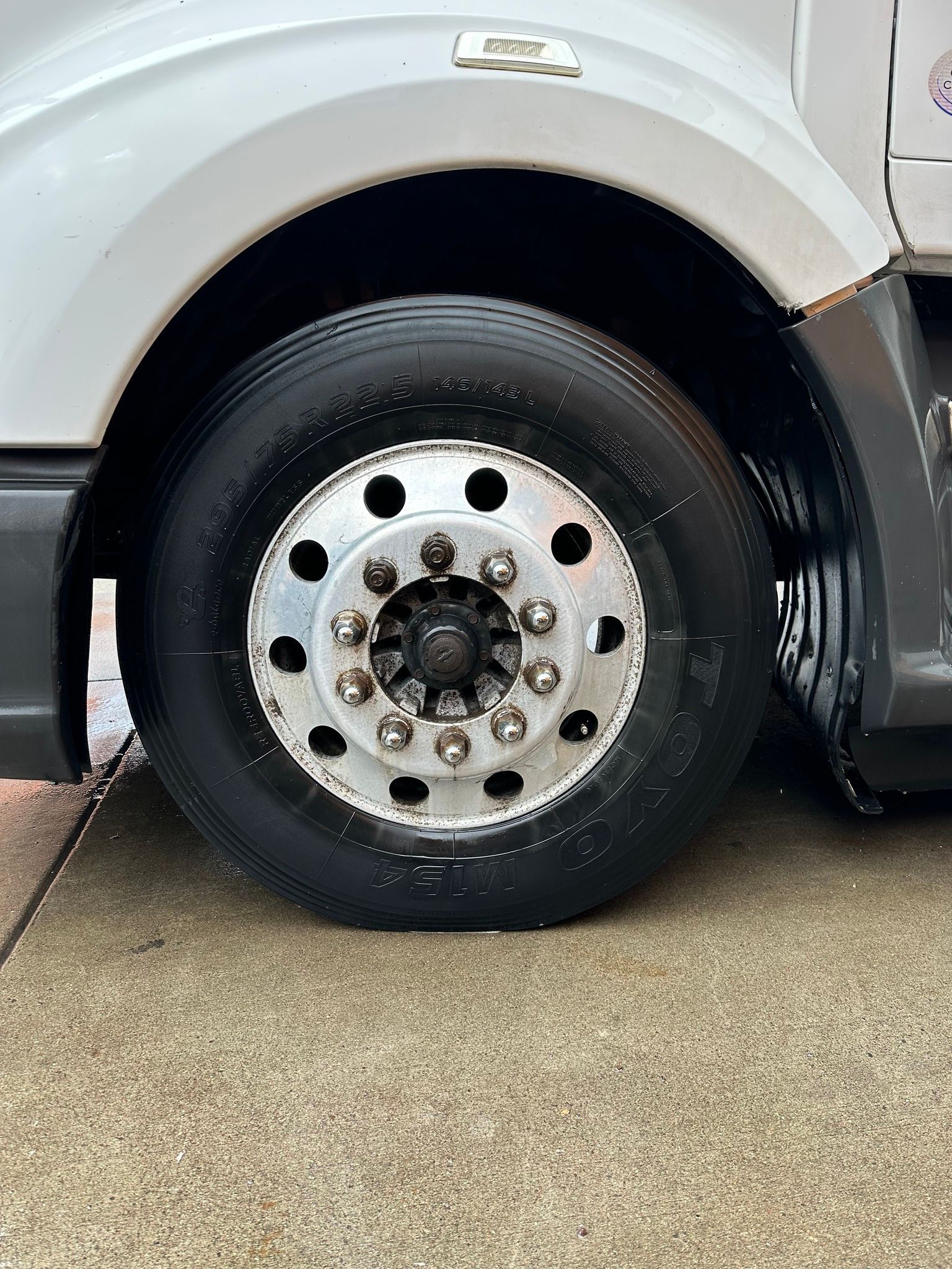 A close-up of a flat tire on a truck, showing the wheel, tire, and part of the truck's body. The truck is parked on a paved surface.