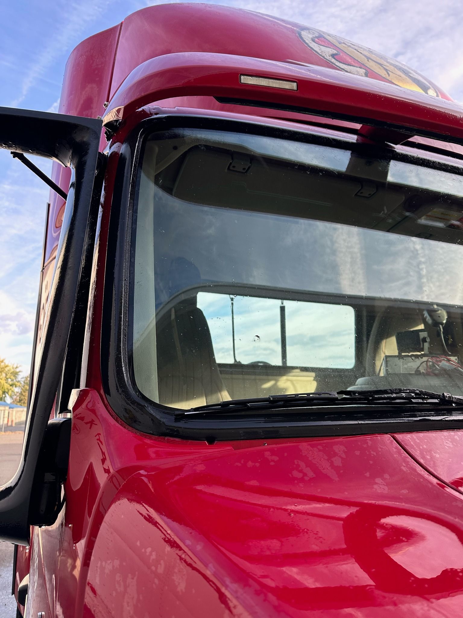 Red semi-truck cab with a large windshield reflecting a blue sky. The side mirror is extended.