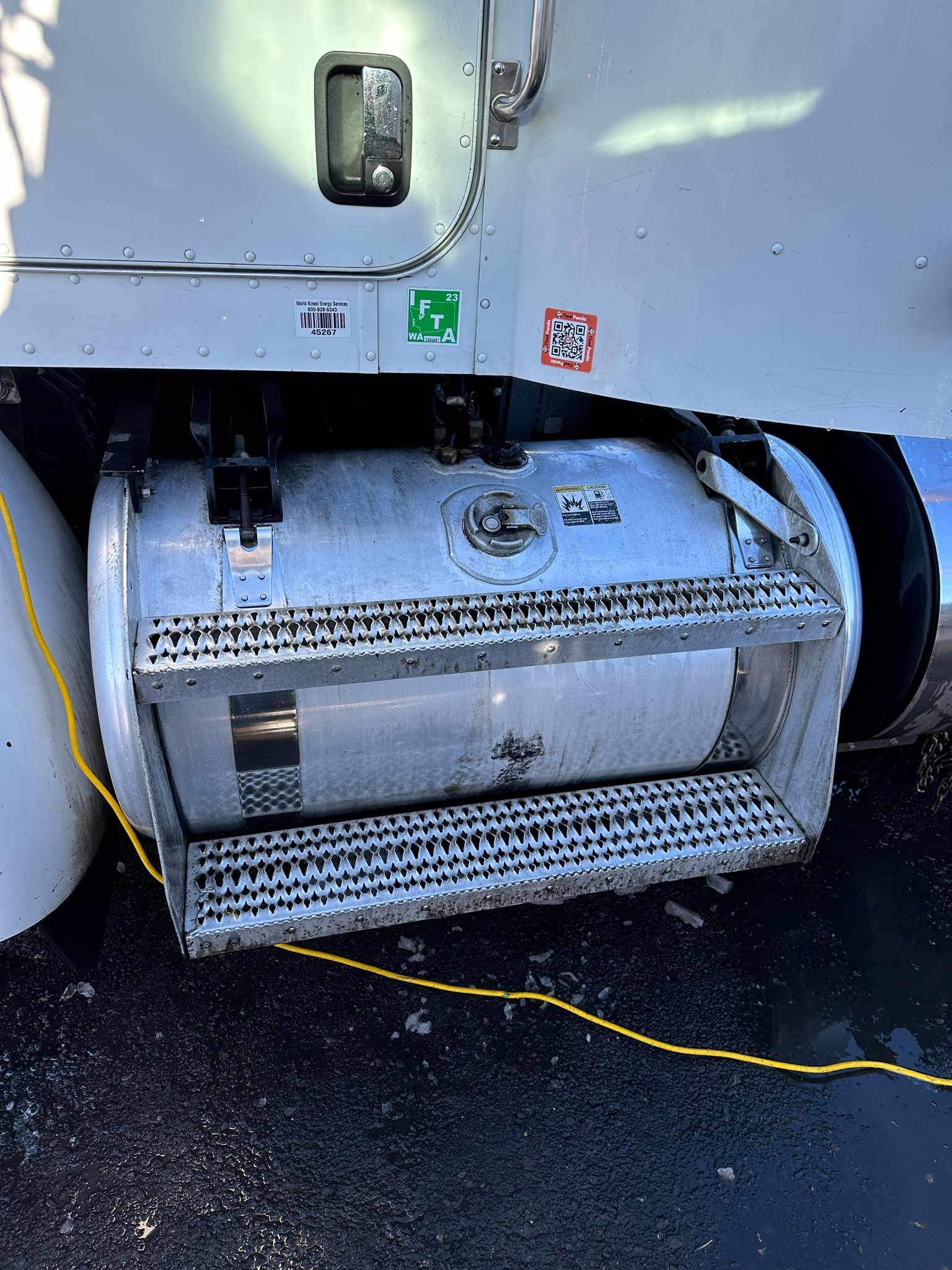 A silver fuel tank with a set of steps attached to a semi-truck, with warning stickers on the truck's body.