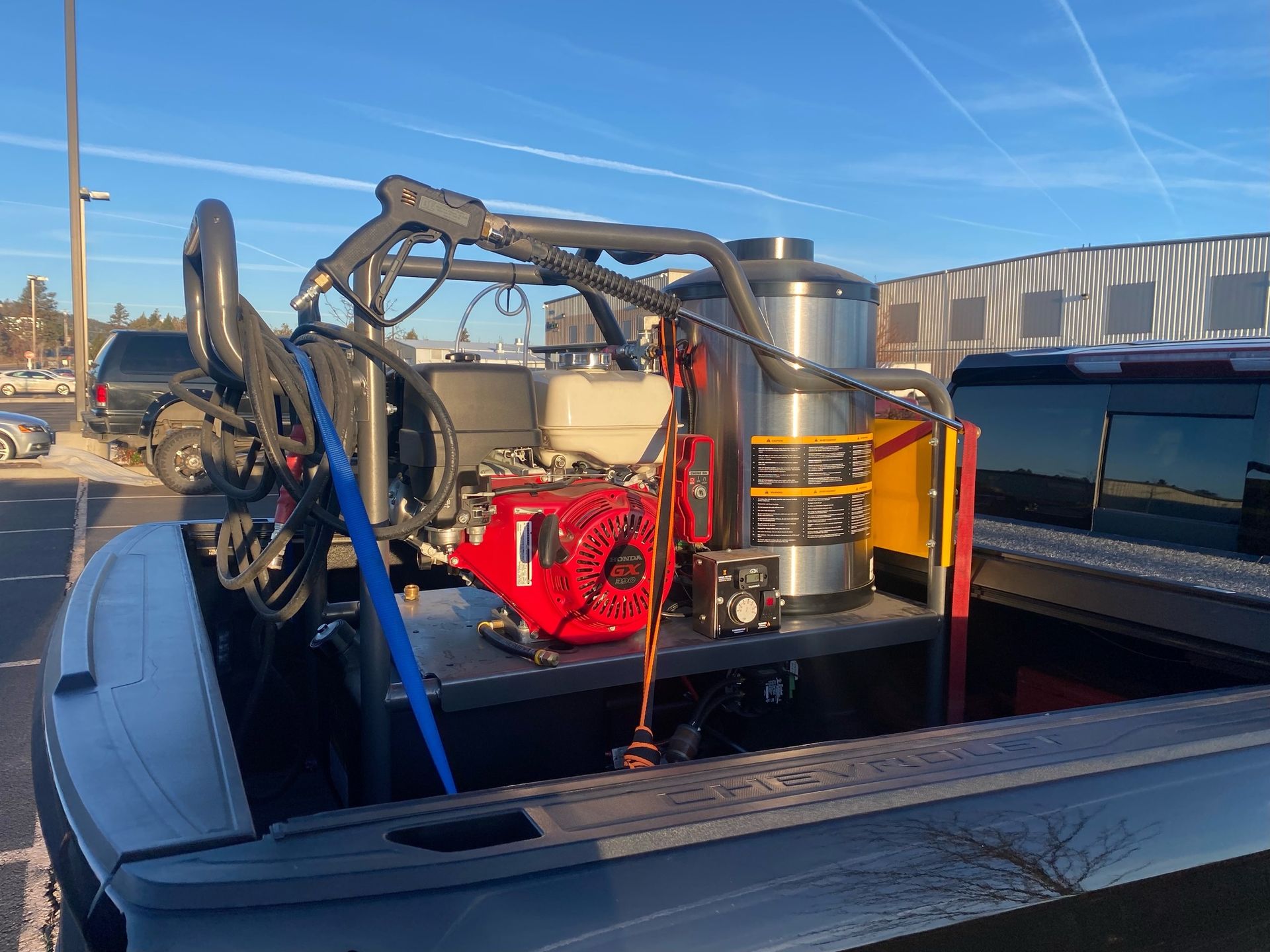 A truck bed fitted with power washing equipment, including a water tank, pumps, and hoses, is parked outdoors on a sunny day.