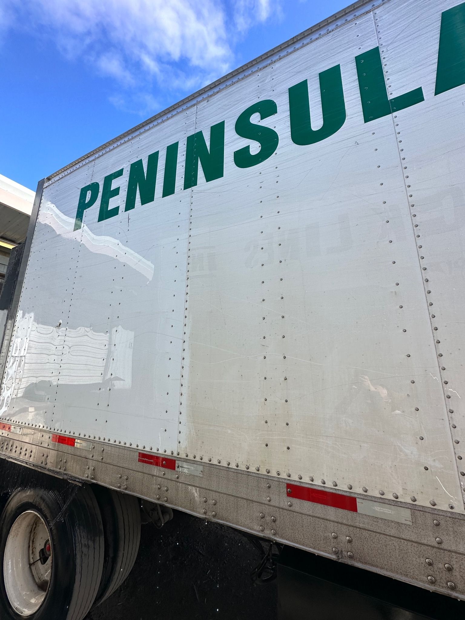 Side of a white Peninsula semi-truck with green lettering and red reflective strips. Blue sky in the background.