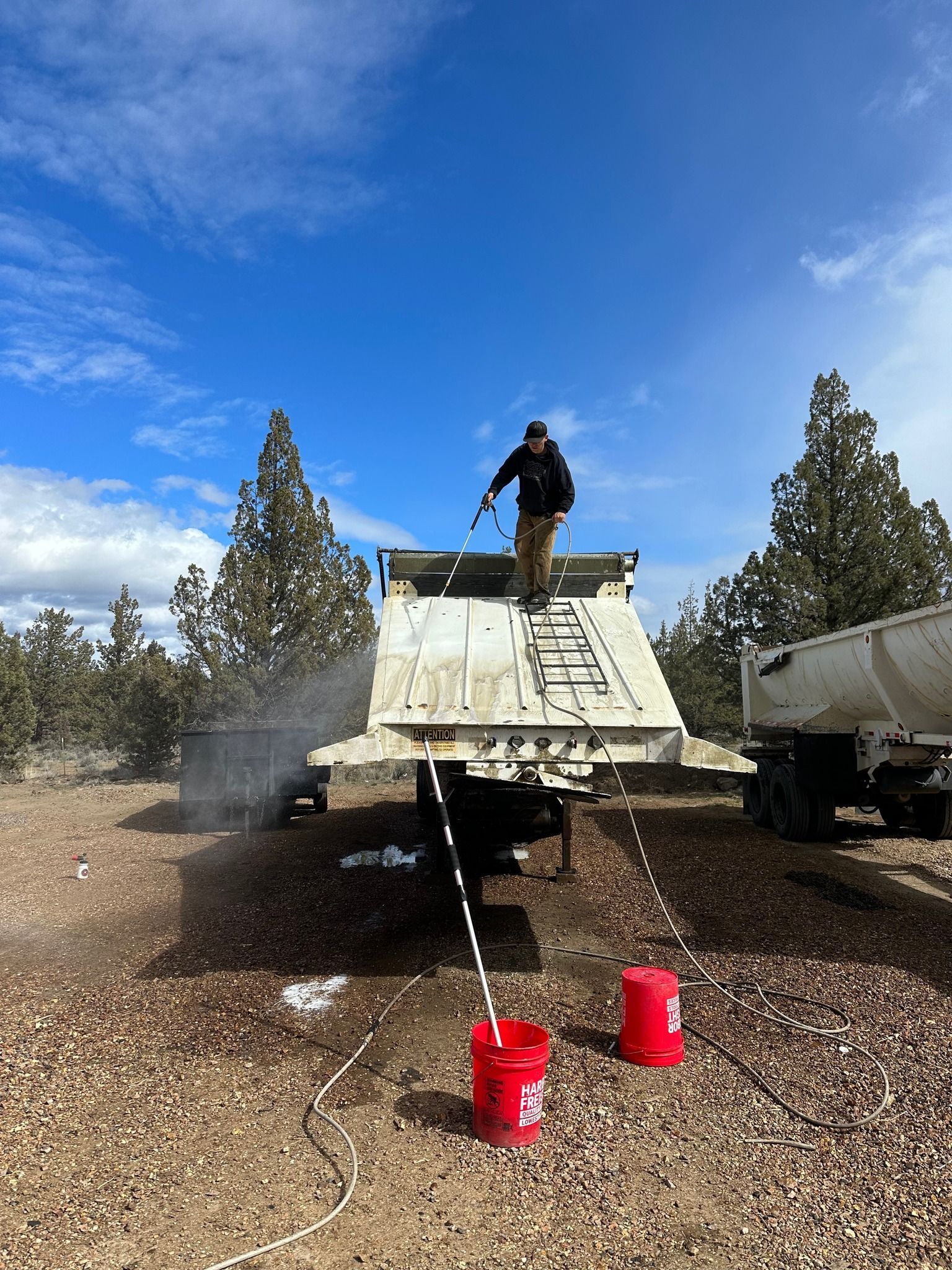 Man washes out a dump truck bed with a pressure washer. Red buckets and a second truck are nearby, under a blue sky.