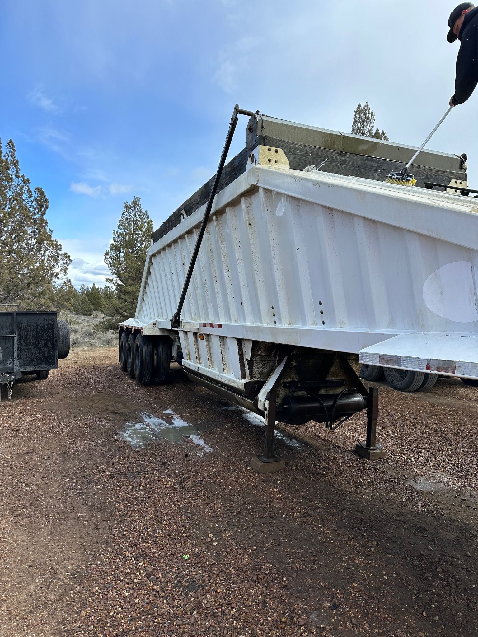 White dump trailer in a dirt lot, tilted upwards, with a person reaching towards it. Blue sky, dry vegetation in background.