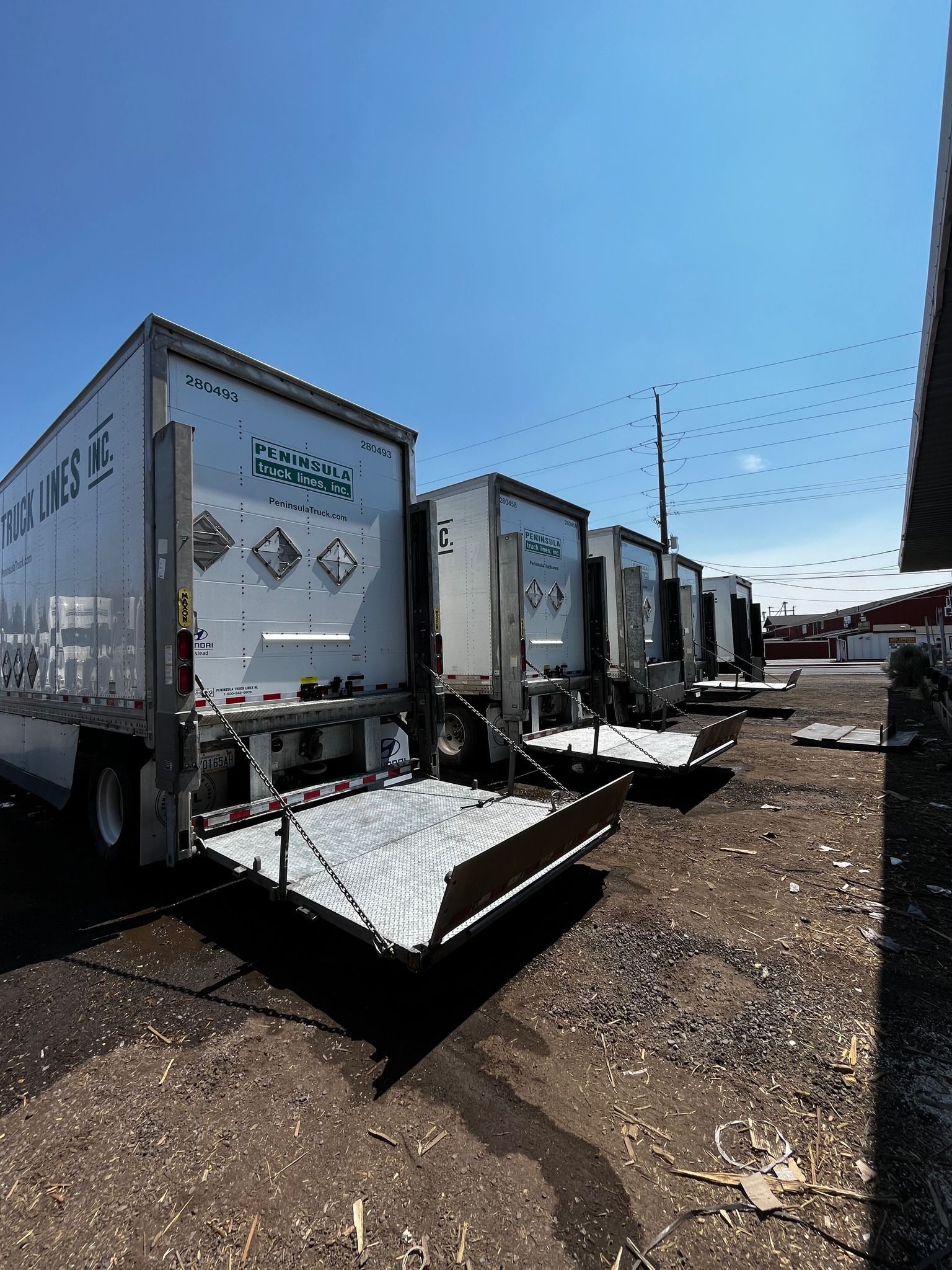 Several white semi-truck trailers parked on a gravel lot, with open liftgates. Blue sky.