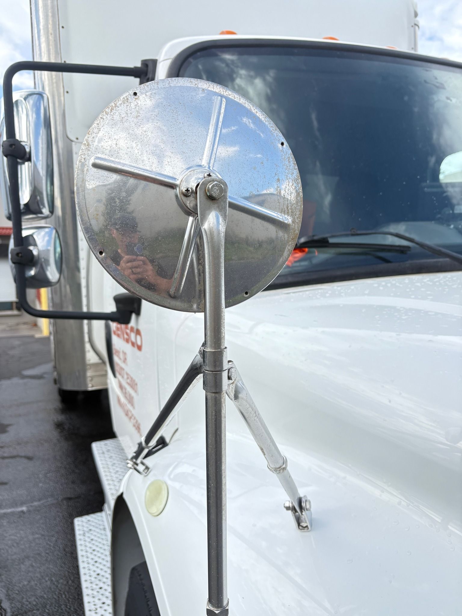 Close-up of a truck's side mirror, showing a large, round, convex mirror with raindrops on it, mounted on a shiny metal arm on the truck's white body.