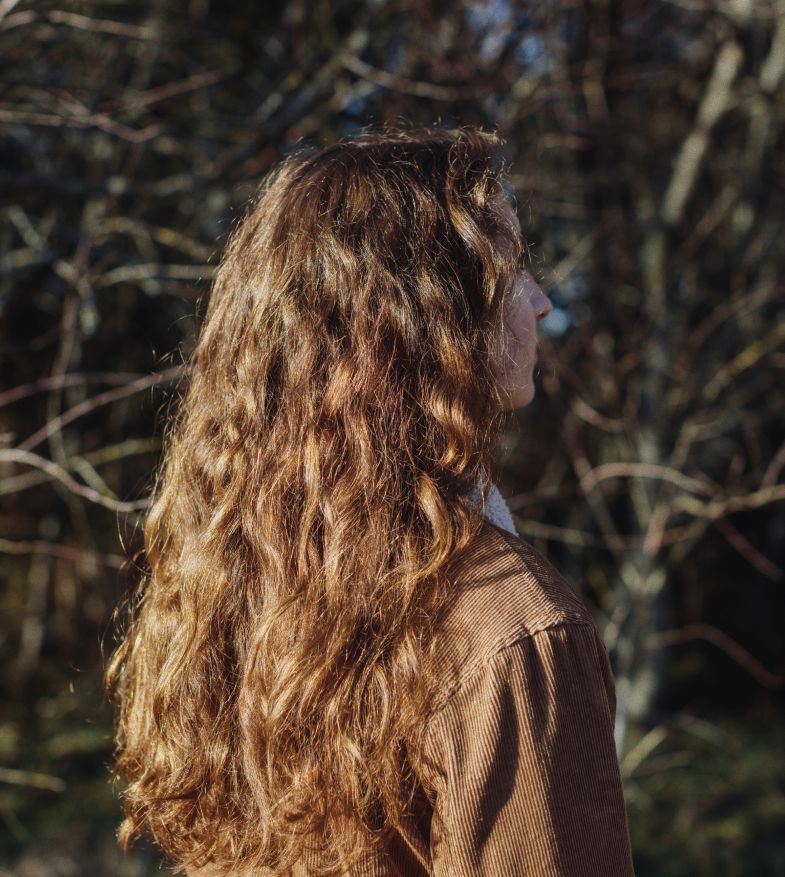 Woman with long, wavy brown hair, outdoors in front of bare tree branches, wearing brown coat, side profile.