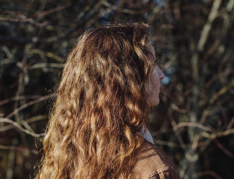 Woman with long, wavy brown hair, outdoors in front of bare tree branches, wearing brown coat, side profile.