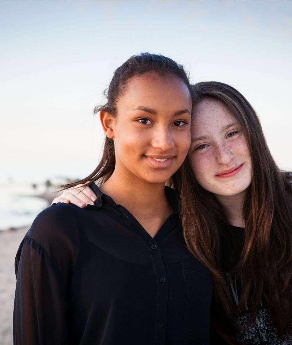 Two smiling young women embrace outdoors. One has dark skin, the other, fair skin and freckles.