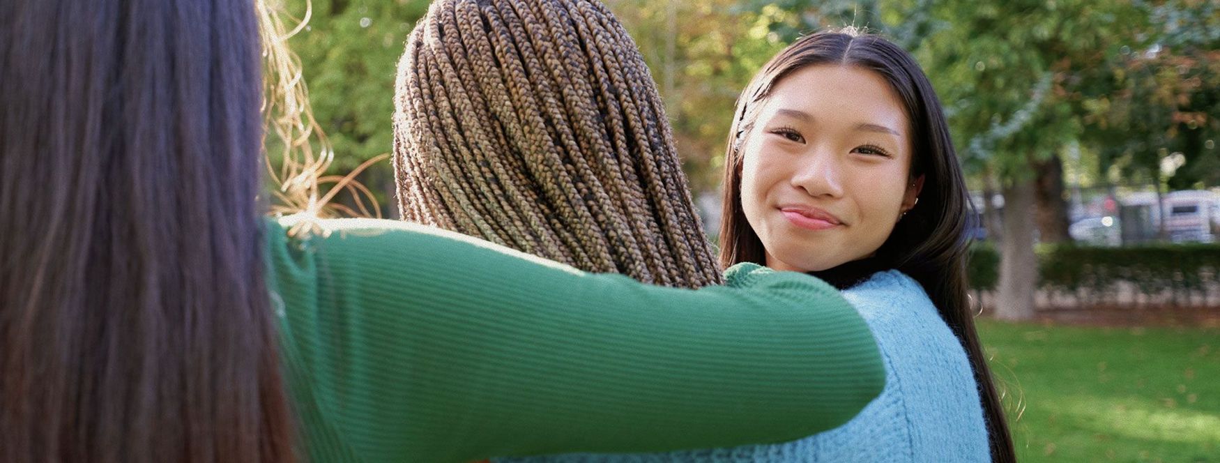 Three people embracing outdoors; one smiling at the camera, sunlight, green grass, and trees.