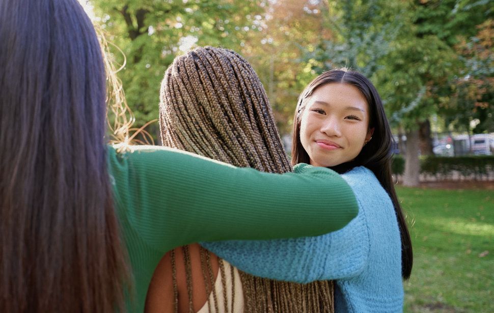 Three people embracing outdoors; one smiling at the camera, sunlight, green grass, and trees.