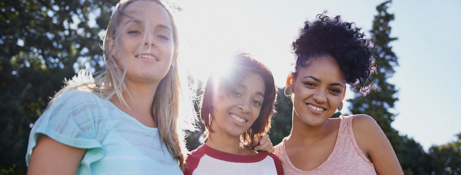 Three young people smiling at the camera outdoors, arms around each other. Sun is shining.