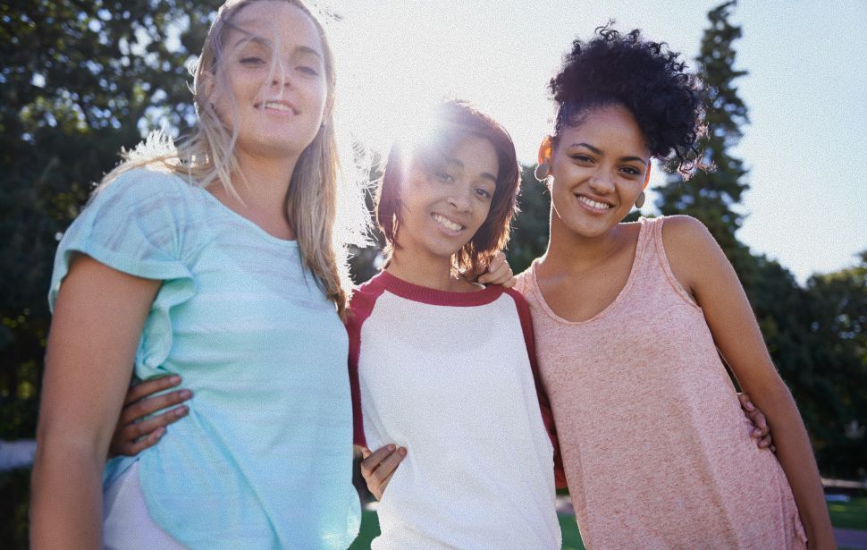Three young people smiling at the camera outdoors, arms around each other. Sun is shining.