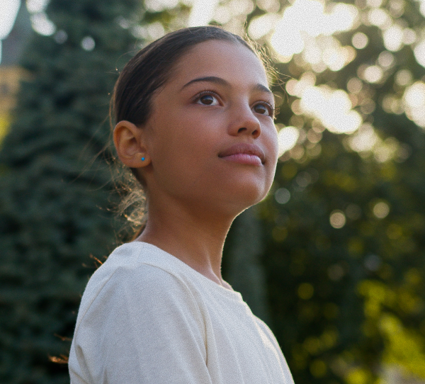 Young person looking up, outdoors. Soft sunlight, trees in background.