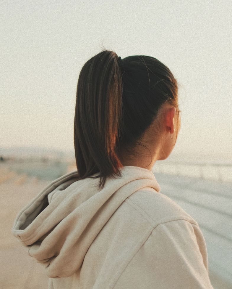 Woman in beige hoodie, hair in a ponytail, looking out at a body of water during the day.