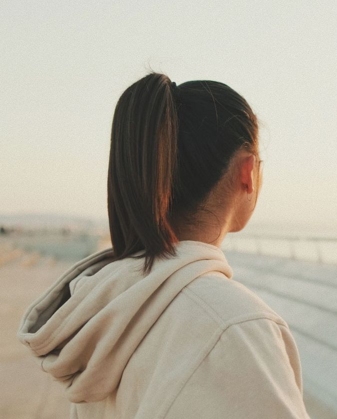 Woman in beige hoodie, hair in a ponytail, looking out at a body of water during the day.