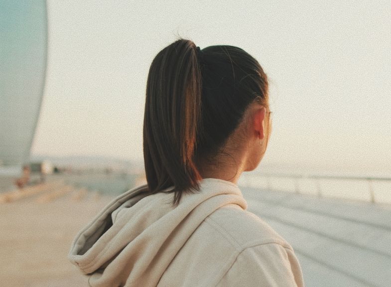 Woman in beige hoodie, hair in a ponytail, looking out at a body of water during the day.
