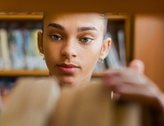 Young person looking at books on a shelf; indoor library setting.