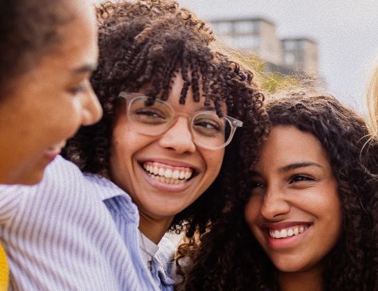 Three smiling people embracing outdoors, one wearing glasses and a light blue shirt.
