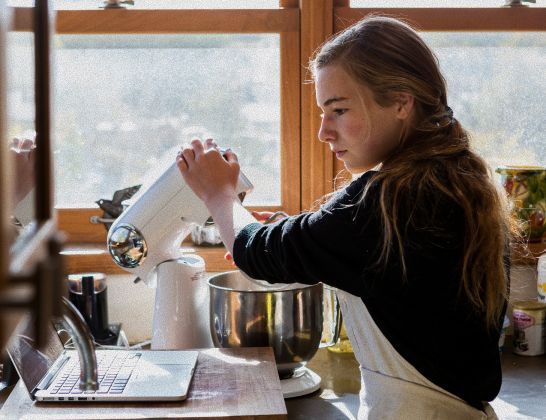 Person pouring into a mixer in a sunlit kitchen. Wearing apron, long hair pulled back.