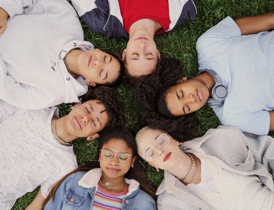 Six people lying in a circle on grass with eyes closed.