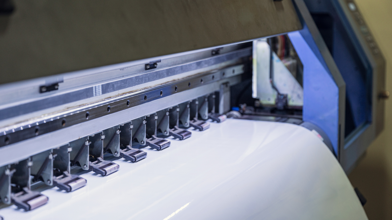 Large format printer with multiple print heads dispensing ink onto a white roll of paper.