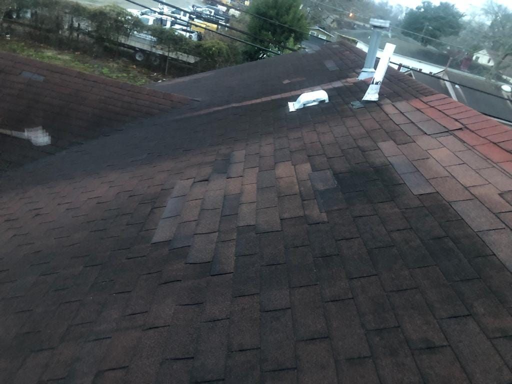 Dark brown asphalt shingle roof with missing and damaged shingles, a metal chimney, and a street in the background.