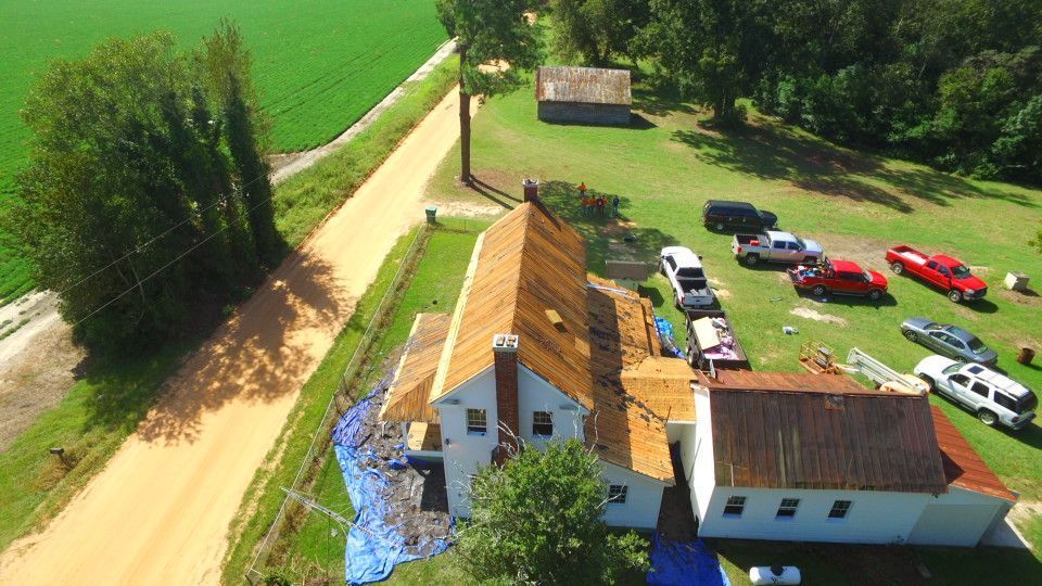 Aerial view of a house with a partially replaced roof, surrounded by cars and farmland.