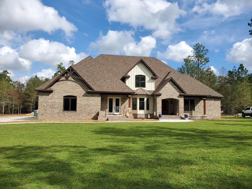 A large, brick house with a brown roof and white trim sits on a green lawn under a blue sky with clouds.