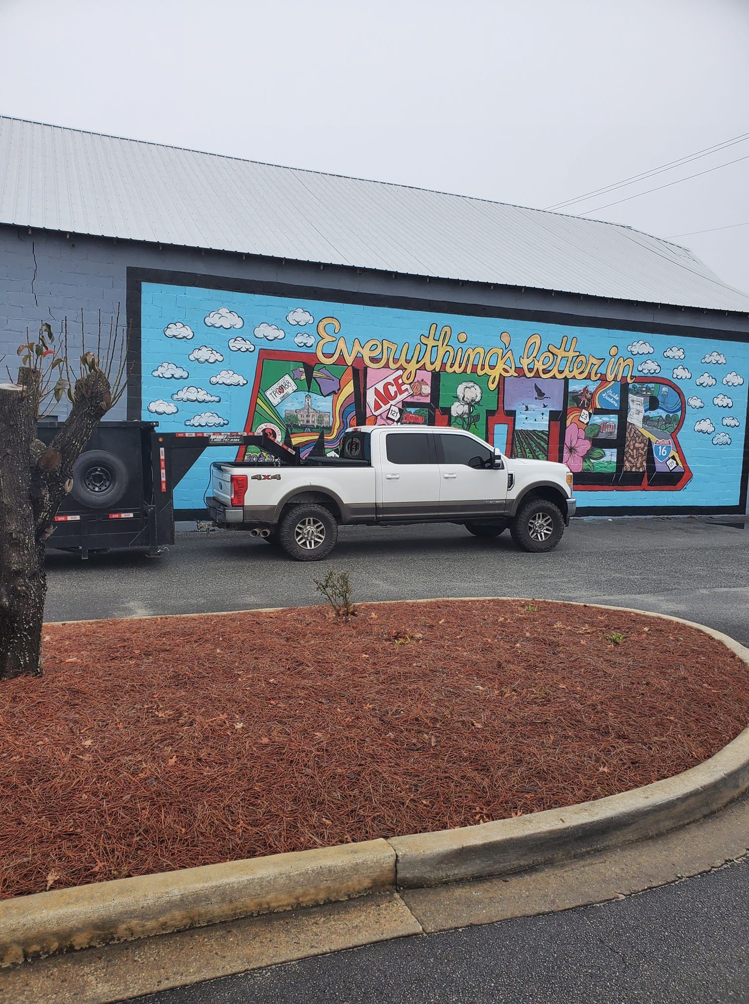White truck with trailer parked in front of a building with a colorful mural. Truck bed holds motorcycles.