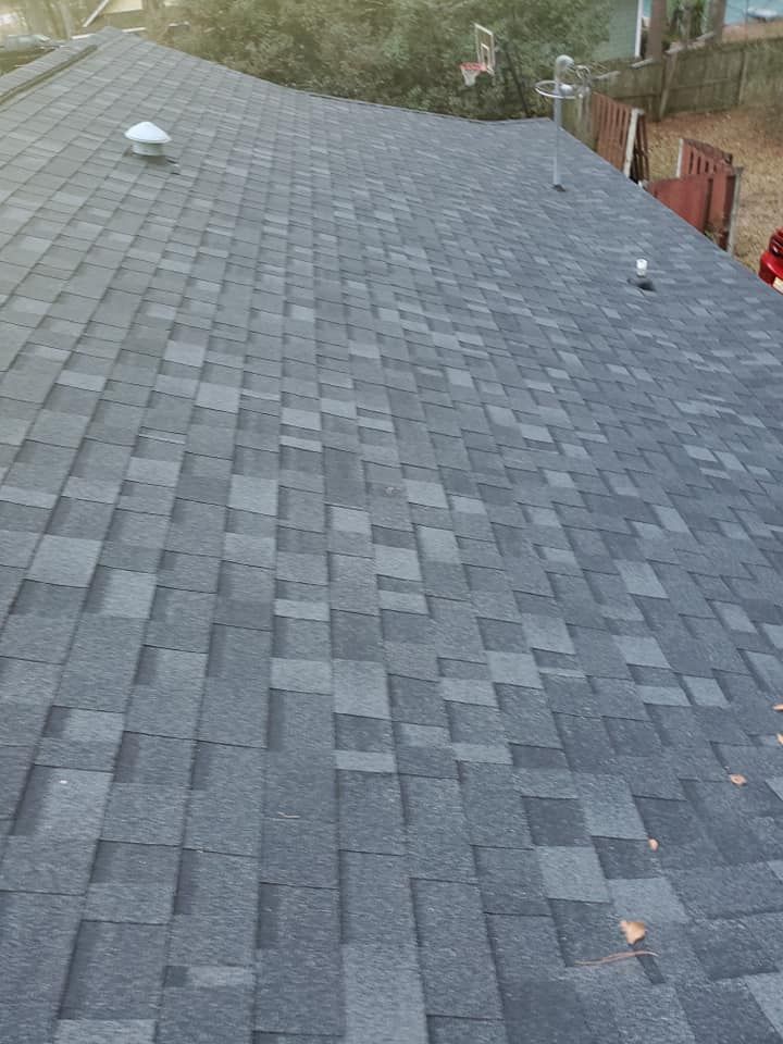 View of a shingled roof with varying shades of gray. Two vents are visible; foliage and a fence are in the background.