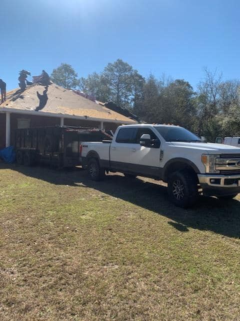 White pickup truck with a trailer parked in front of a house where roofers are working on the roof on a sunny day.