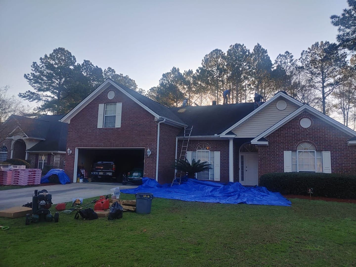 A brick house with a new roof, covered in blue tarps. Landscaping equipment and a ladder are on the lawn.