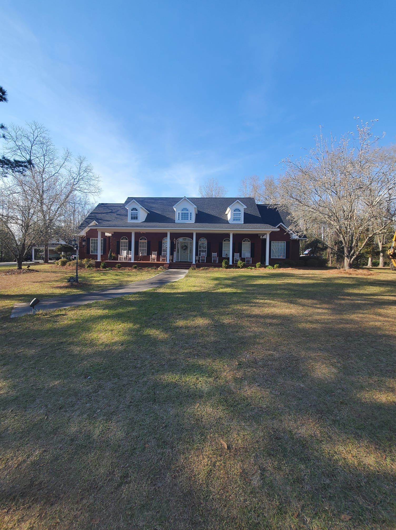 A brick, single-story house with a long porch, white columns, and dormer windows sits on a large lawn under a blue sky.