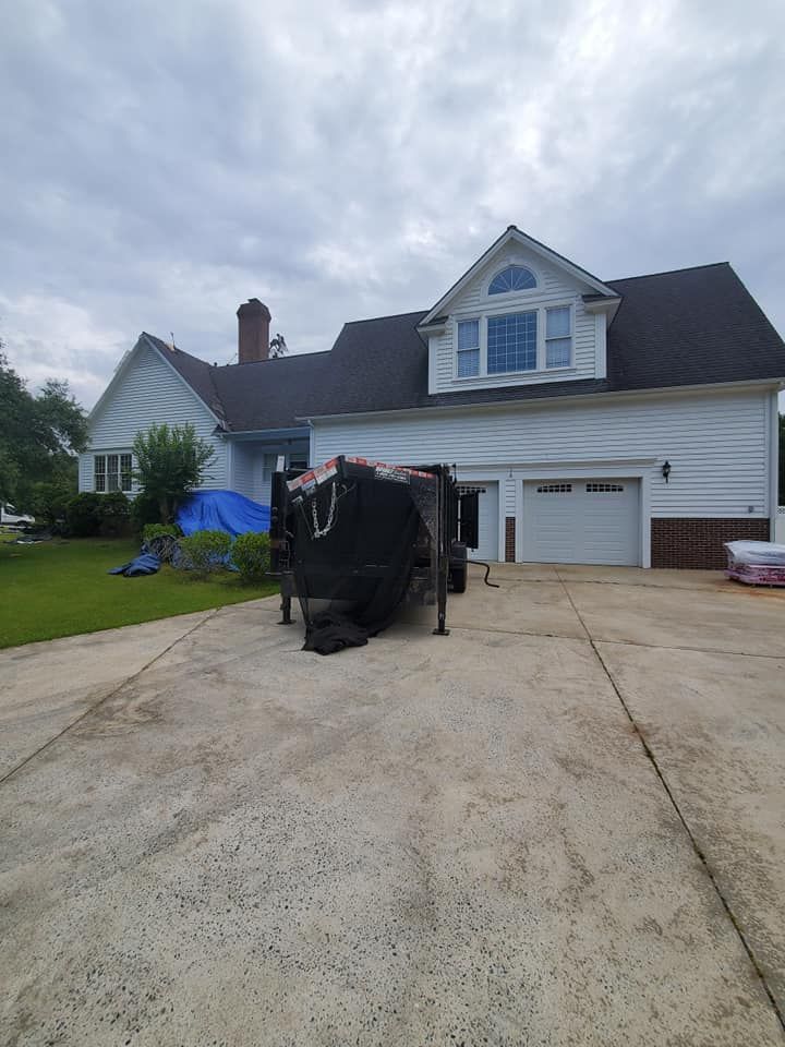 A white house with a black roof, a covered trailer parked in the driveway, and a cloudy sky overhead.